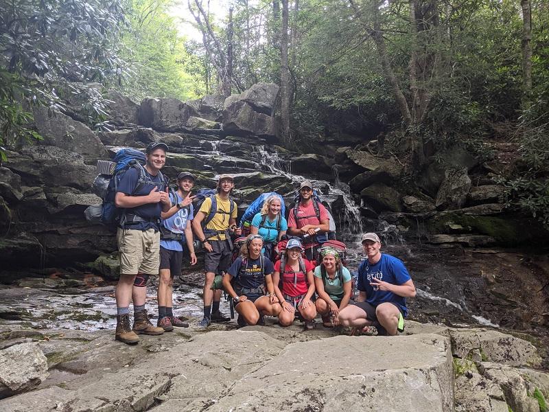 Group of students smiling in front of a waterfall they hiked to on an Outdoor trip.